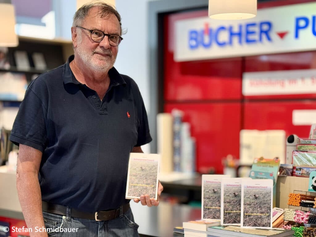 Mann mit Brille und Bart hält in einem Buchladen ein Buch in der Hand; im Hintergrund sind Bücherregale und das teilweise sichtbare Schild „BÜCHER“ zu sehen.