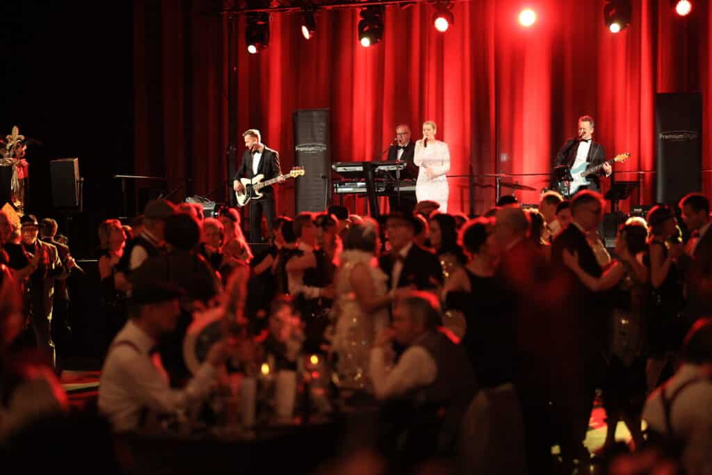 Eine Liveband spielt vor roten Vorhängen auf einer beleuchteten Bühne, während Gäste im Vordergrund feiern und tanzen.
