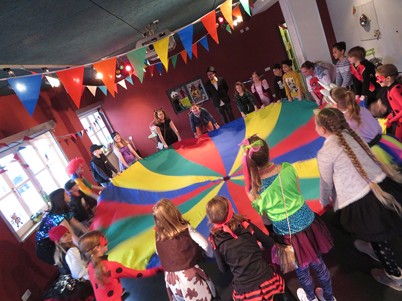 Kinder und Jugendliche spielen beim Kinderfasching mit einem bunten Schwungtuch in einem festlich geschmückten Raum.