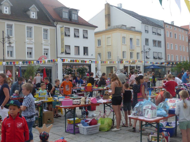 Kinder und Erwachsene stöbern an Verkaufsständen eines Kinderflohmarkts auf einem belebten Platz in Deggendorf.