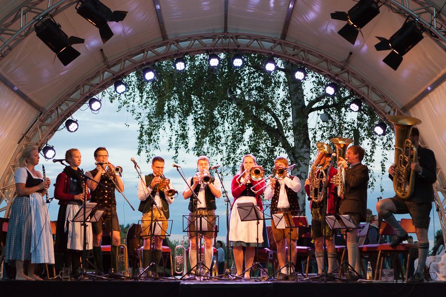 Blasorchester auf einer Bühne im Festzelt Musikerinnen und Musiker eines Blasorchesters spielen unter einer überdachten Bühne vor einem Baumensemble.