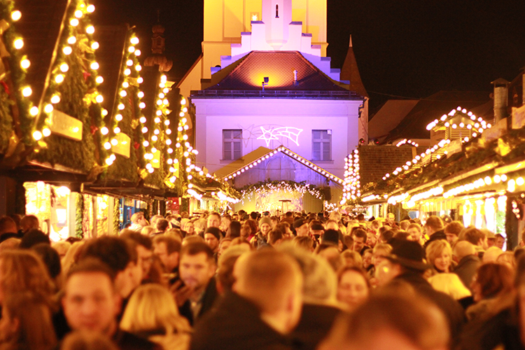 Dicht gedrängte Menschen auf einem weihnachtlich beleuchteten Christkindlmarkt vor einem festlich angestrahlten Gebäude.