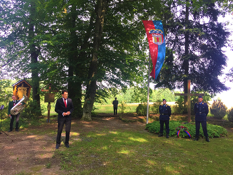 Gedenkveranstaltung am Gefallenenhain Mehrere Personen versammeln sich an einer Gedenkstätte im Grünen; neben einem Fahnenmast mit Wappen stehen ein Redner und uniformierte Teilnehmer.