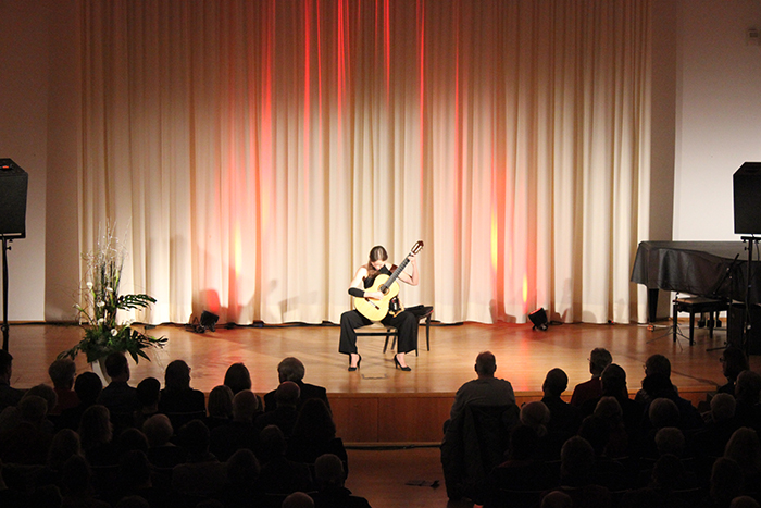 Gitarrenkonzert im Veranstaltungssaal Eine sitzende Musikerin spielt auf einer Bühne vor einem Publikum klassische Gitarre. Hinter ihr hängen helle Vorhänge mit roter und orangefarbener Beleuchtung; seitlich stehen Lautsprecher und ein Flügel.