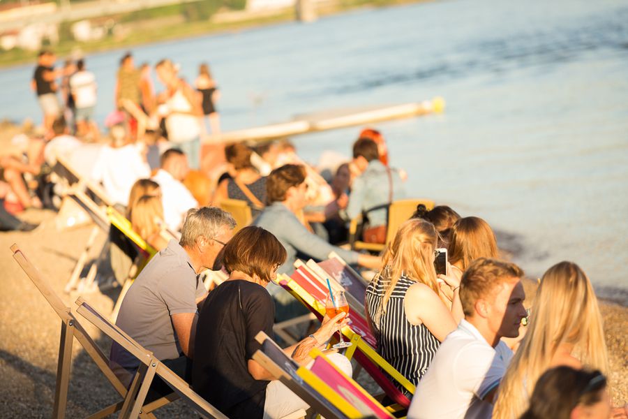 Ruderverein Deggendorf beim Sommerfest an der Donau Menschen sitzen bei einer Veranstaltung auf Liegestühlen am Donauufer; im Vordergrund liegen farbenfrohe Ruder und Boote.
