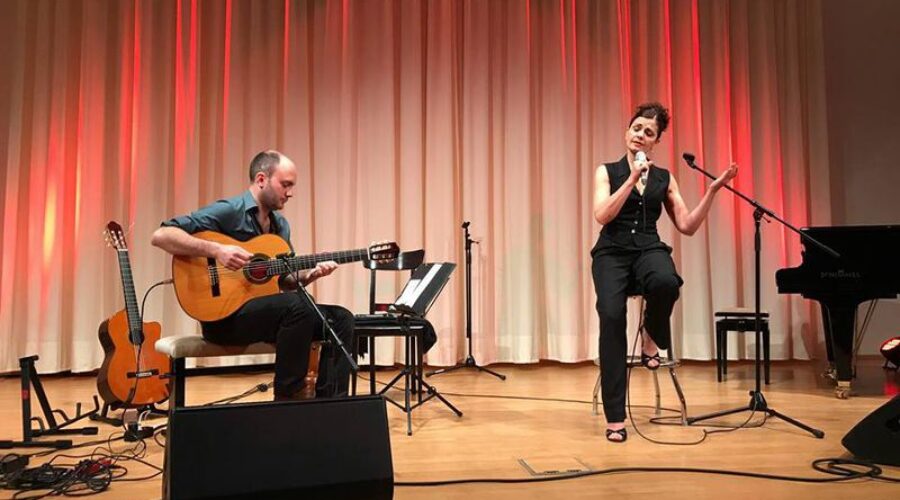 Konzert mit Sängerin und Gitarrist auf der Bühne Sängerin mit Mikrofon und Gitarrist mit Akustikgitarre bei einem Konzert auf einer beleuchteten Bühne; rechts steht ein Flügel.