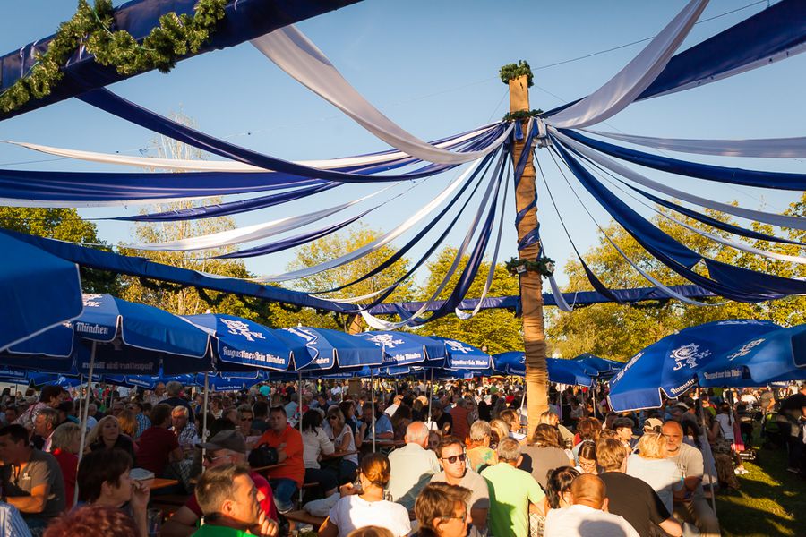 Sommerfest des Vereins in Deggendorf 2003 Dicht besuchtes Vereinsfest mit Gästen unter einem mit blau-weißen Stoffbahnen geschmückten Maibaum; im Hintergrund stehen blaue Sonnenschirme und Bäume.