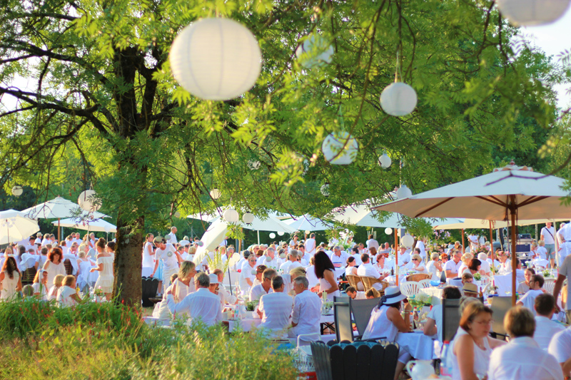 Gäste in weißer Kleidung sitzen bei einem sommerlichen Dinner im Grünen an gedeckten Tischen; über ihnen hängen weiße Papierlaternen.
