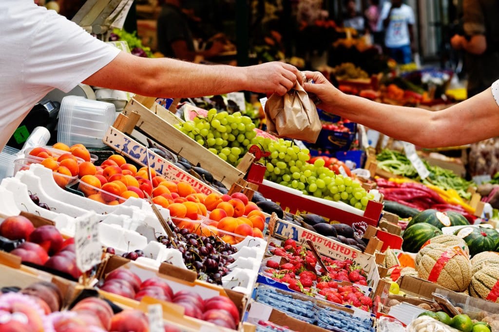 Hände reichen sich an einem Wochenmarkt eine Papiertüte über Auslagen mit Obst und Gemüse.
