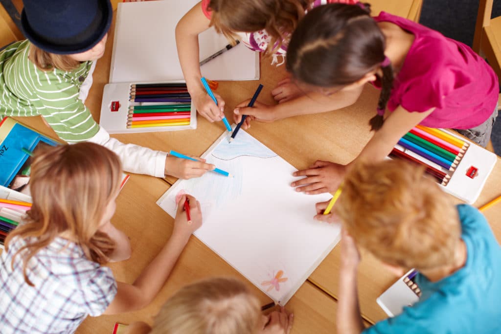 Mehrere Kinder gestalten gemeinsam mit Buntstiften ein großes Plakat an einem Tisch.