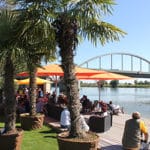 Tourismusveranstaltung am Wasser Menschen sitzen unter Sonnenschirmen auf einer Terrasse am Wasser; Palmen und eine Brücke sind im Hintergrund zu sehen.