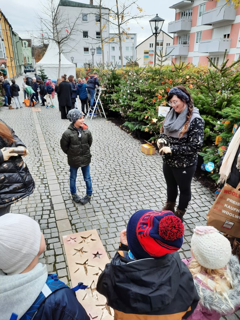 Kinder und Erwachsene stehen auf einem gepflasterten Platz neben geschmückten Weihnachtsbäumen; eine Person zeigt auf einen Sternenwegweiser zur Kinderweihnacht.