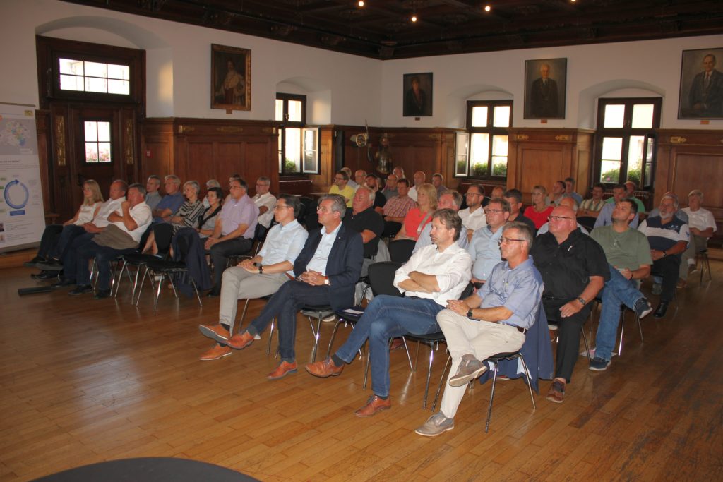Informationsveranstaltung im Auditorium des Interkom-Rats Sitzreihen mit zahlreichen Teilnehmenden bei einer Informationsveranstaltung in einem historischen holzgetäfelten Saal; links steht eine Präsentationstafel.