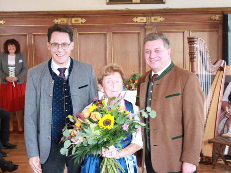 Ehrung mit Blumenstrauß im festlichen Rahmen Eine ältere Frau hält einen großen Blumenstrauß und steht zwischen zwei lächelnden Männern in Tracht; im Hintergrund sind eine Harfe und festlich gestaltete Holzvertäfelungen zu sehen.
