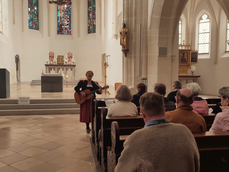 Konzert in einer Kirche in Deggendorf Eine Musikerin spielt vor dem Altar einer Kirche Gitarre und singt, während Besucherinnen und Besucher in den Kirchenbänken zuhören.