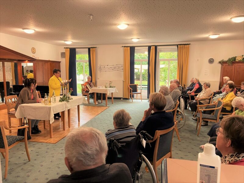 M23 16: Veranstaltung mit Seniorinnen und Senioren Seniorinnen und Senioren sitzen in einem hellen Veranstaltungsraum, während eine Frau an einem gedeckten Tisch spricht.
