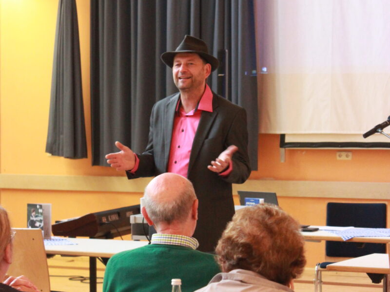 Vortrag eines Referenten vor Publikum Ein Referent mit Hut spricht in einem Saal vor mehreren sitzenden Personen.