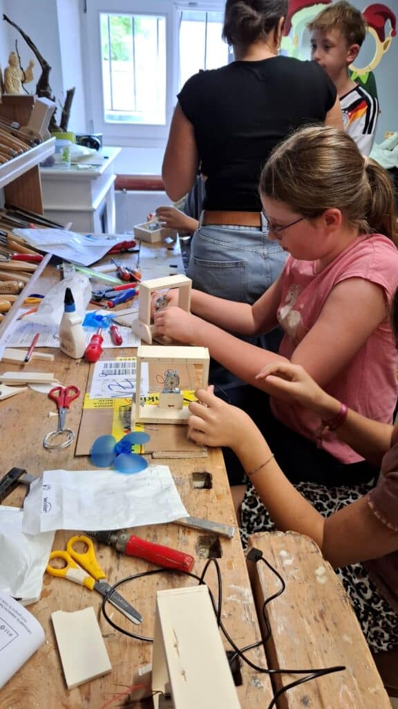Kinder und Jugendliche bauen an einem Werkstatttisch einen kleinen Tischventilator aus Holz und Bastelmaterialien.