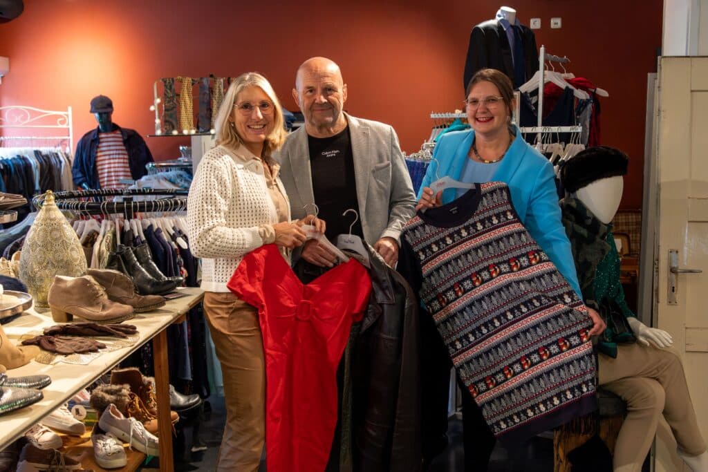 Drei Personen präsentieren Kleidung und Accessoires zwischen Kleiderständern bei einer Kleidertauschparty.
