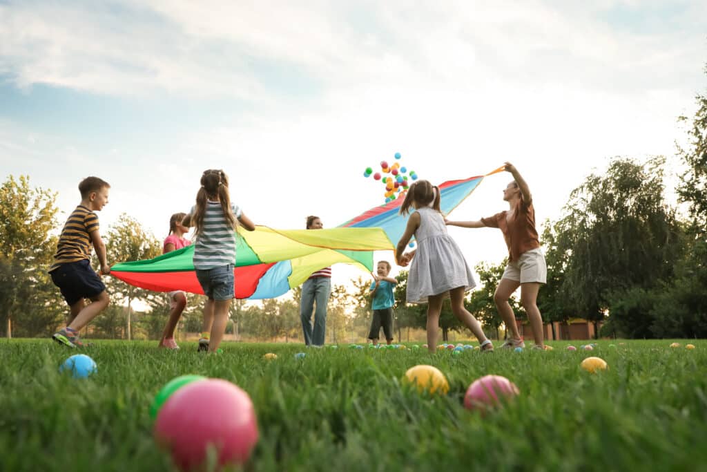 Kinder und Jugendliche spielen auf einer Wiese mit einem bunten Schwungtuch; im Vordergrund liegen farbige Bälle.