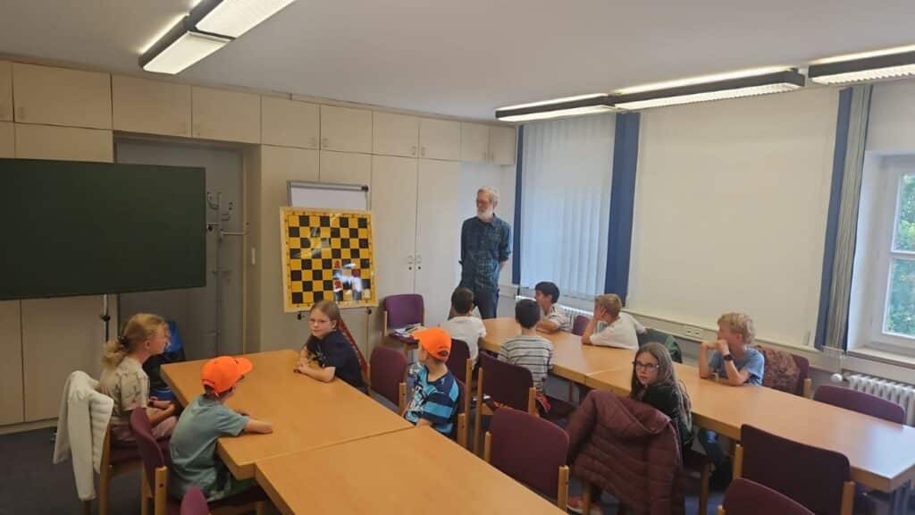 Kinder und Erwachsene sitzen in einem Unterrichtsraum an Tischen; im Hintergrund steht ein Schachbrett.