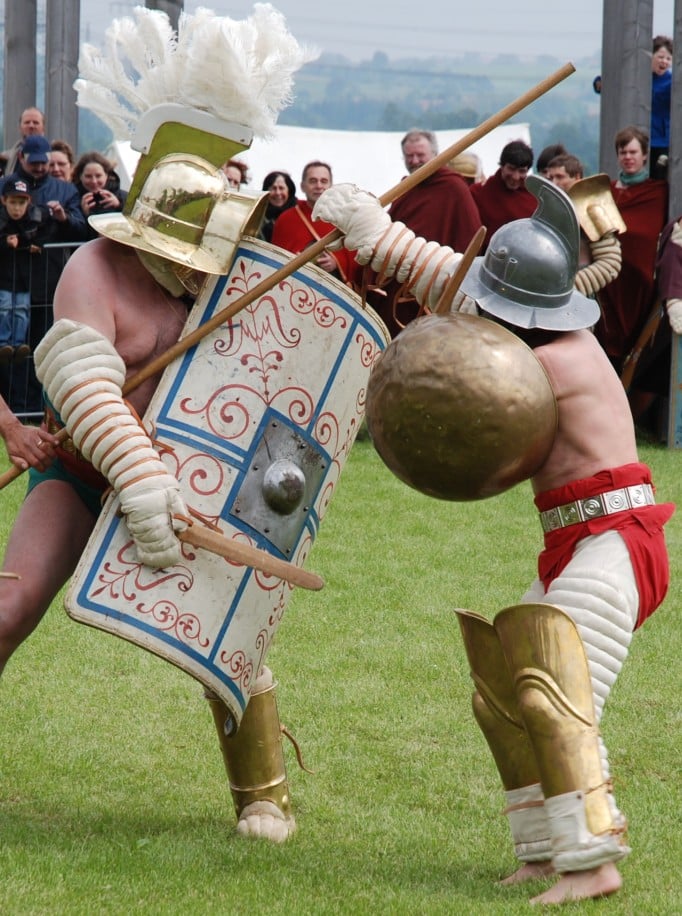 Zwei kostümierte Gladiatoren kämpfen mit Schilden, Schwertern und Speeren auf einer Wiese, während Zuschauer zusehen.