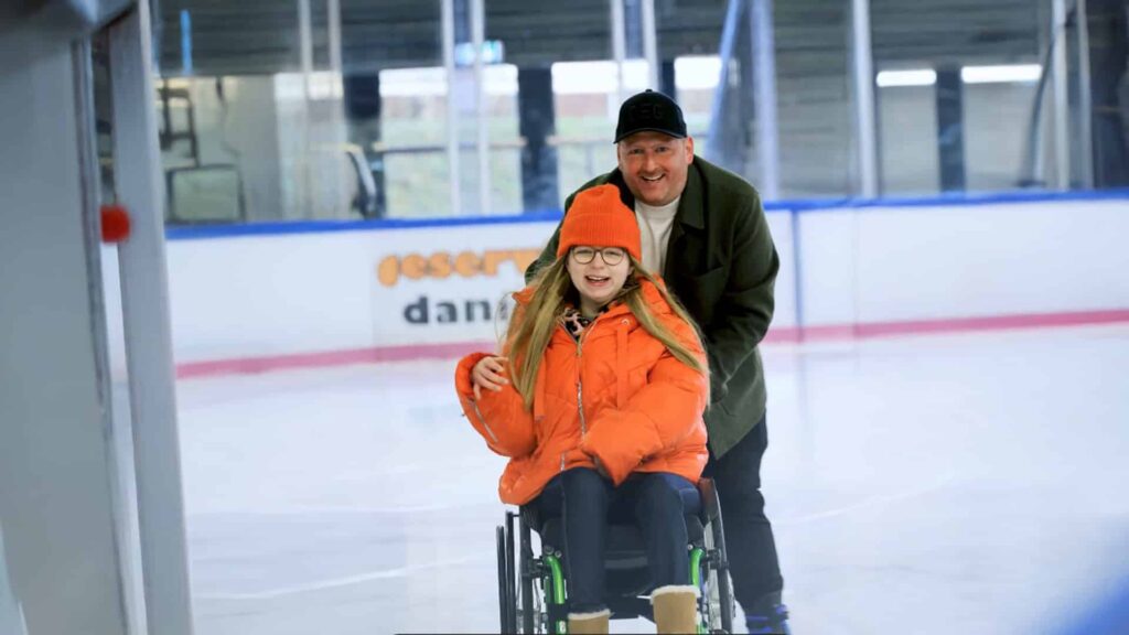 Eine Rollstuhlfahrerin in orangefarbener Jacke fährt mit einer Begleitperson über eine Eisbahn.