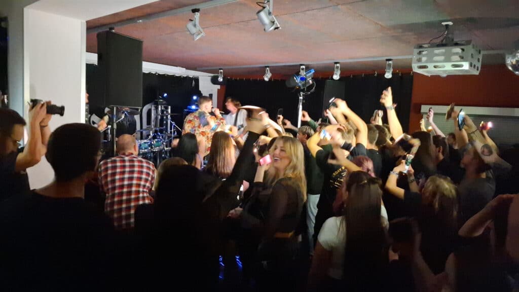 Konzert in einem kleinen Saal: Eine Band spielt auf der Bühne, während zahlreiche Menschen im Publikum tanzen und ihre Arme heben.