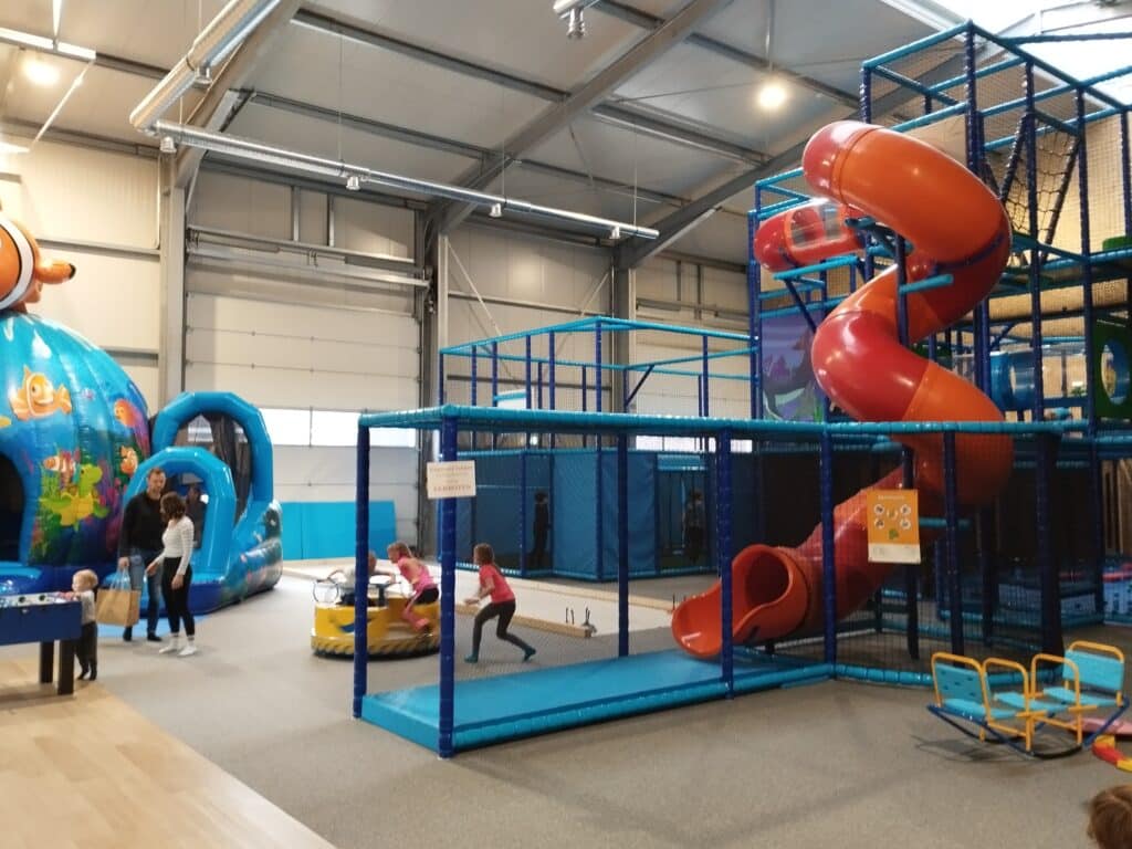Kinder spielen auf Hüpfburgen, in einem Kletterbereich und auf einer Rutsche in einer großen Indoor-Spielhalle.