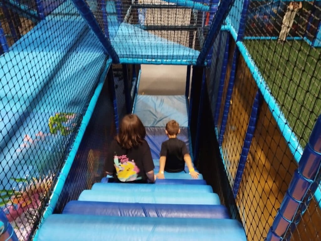 4You-Gaudium: Indoor-Spielbereich in Deggendorf, Saison 2024 Zwei Kinder klettern in einem blau gepolsterten Indoor-Spielbereich eine Treppe hinunter; Netze und Spielgeräte umgeben den Parcours.