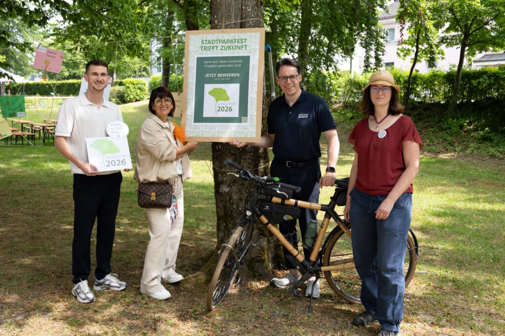 Vier Personen präsentieren im Park ein Plakat und eine Mappe zum Stadtfahrrad-Test 2026 neben einem Fahrrad.