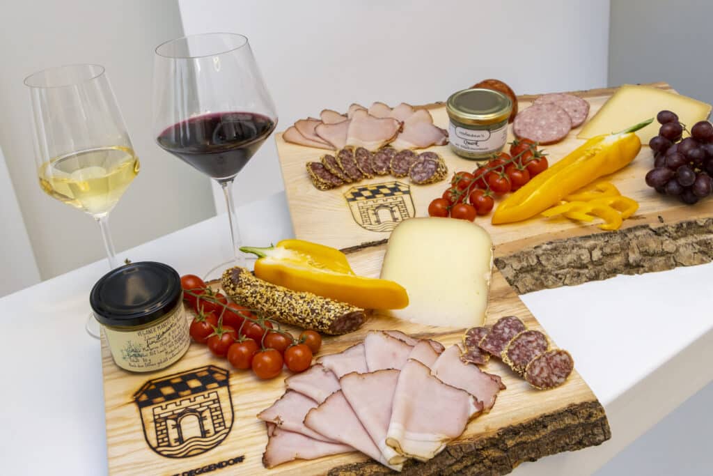 Zwei Gläser mit Weiß- und Rotwein neben Käse, Wurst, Schinken, Tomaten, Paprika, Trauben und Brot auf einem gedeckten Tisch.