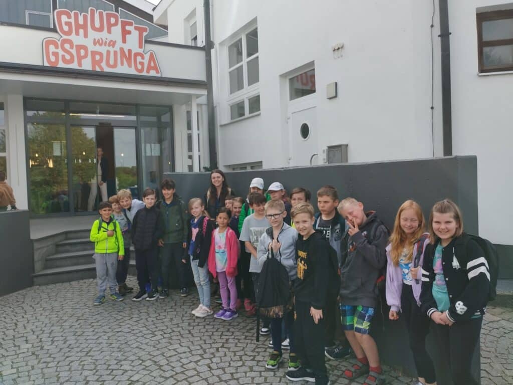 Kinder und Jugendliche stehen vor einem Gebäudeeingang mit dem Schriftzug „GHUPFT WIA GSPRUNGA“.