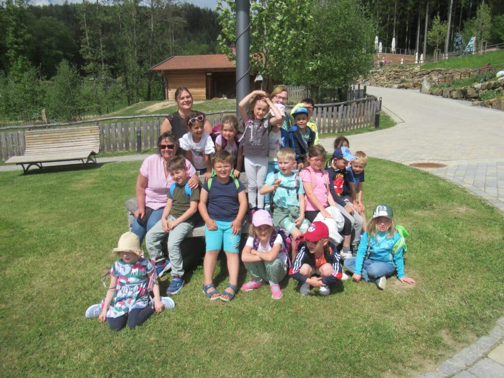 Kindergarten Rettenbach bei der Saisonveranstaltung Edelwies Eine Gruppe von Kindern und erwachsenen Begleitpersonen sitzt und steht auf einer Wiese vor einem Holzgebäude und einem bewaldeten Gelände.