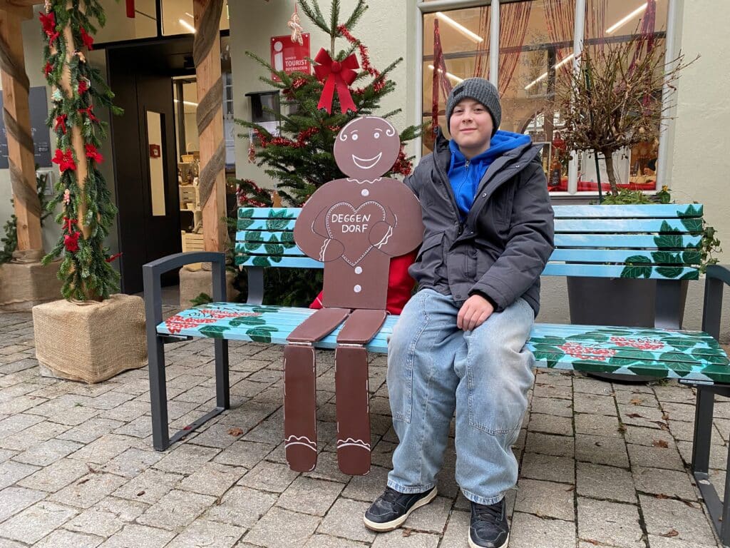 Lebkuchen-Photopoint mit Konstantin und Linus Ein Junge sitzt auf einer weihnachtlich dekorierten Bank neben einer Lebkuchenfigur mit der Aufschrift „DEG?ER DORF“; dahinter stehen ein geschmückter Weihnachtsbaum und ein roter Weihnachtsmann.