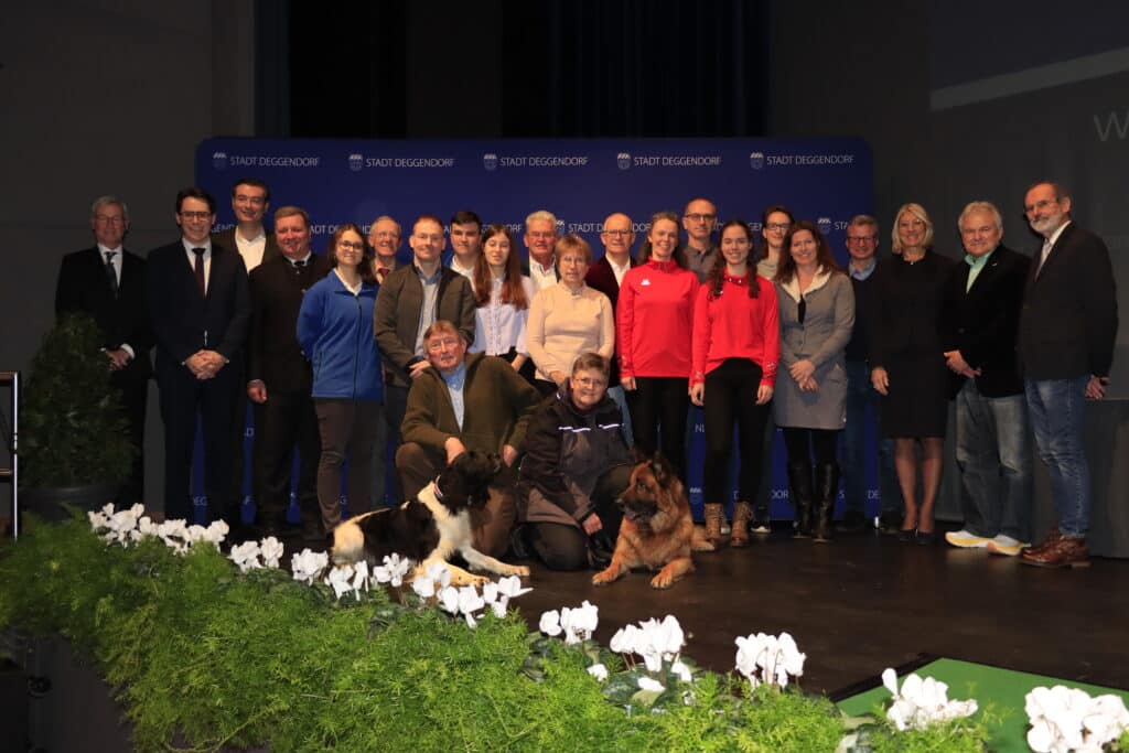 Gruppenfoto bei einer Veranstaltung in Deggendorf Gruppe von Menschen steht auf einer Bühne vor einem Veranstaltungsbanner; davor sitzen zwei Personen mit zwei Hunden, umgeben von weißen Blumen und Grünpflanzen.