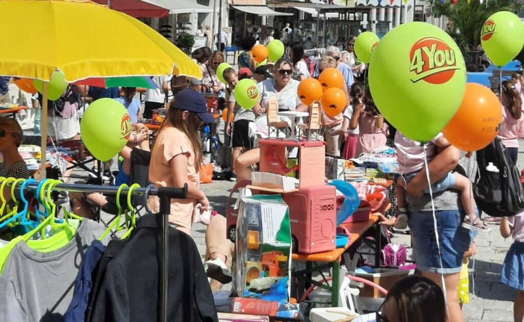 Belebter Kinderflohmarkt unter freiem Himmel mit Kleidung, Spielzeug, Haushaltsgegenständen und Luftballons mit dem Aufdruck „4You“.
