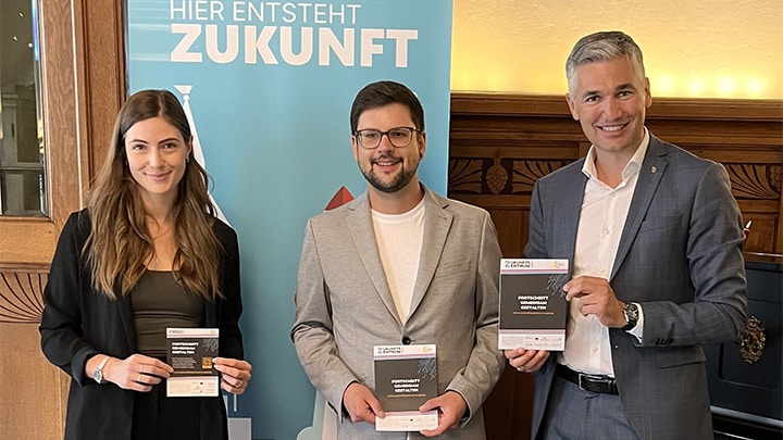 Beratungstag: Drei Personen präsentieren Informationsmaterial Drei Personen stehen in einem holzgetäfelten Raum und halten Informationsmaterial in die Kamera; im Hintergrund steht ein Banner mit der Aufschrift „Hier entsteht Zukunft“.