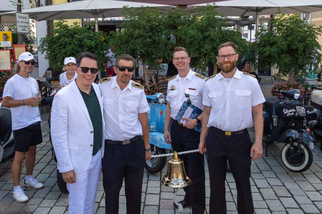 Vier Männer posieren vor einem Motorroller und halten eine goldene Glocke sowie eine gerahmte Auszeichnung.