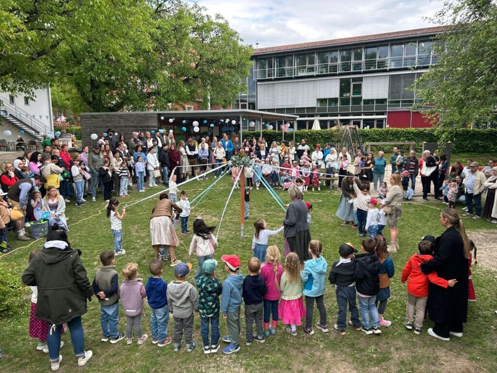 Kinder tanzen und feiern beim Maifest auf einer Wiese, umgeben von Familien und Zuschauenden.
