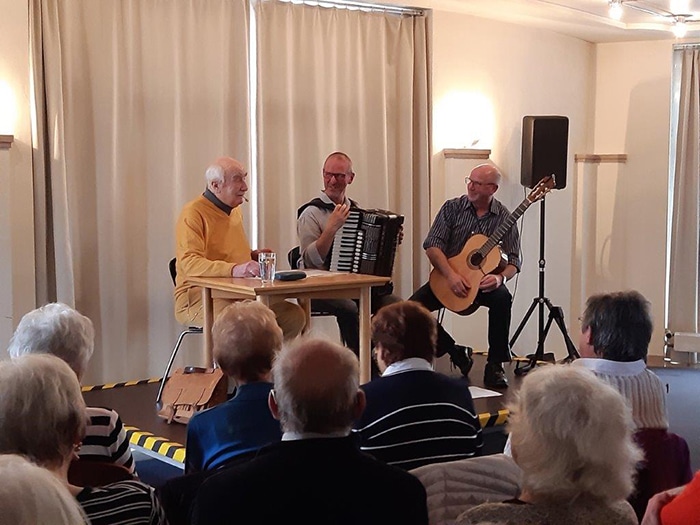 Musiker bei einer Veranstaltung vor Publikum Drei Musiker spielen vor sitzendem Publikum; ein Mann singt mit Akkordeon, ein anderer begleitet ihn auf der Gitarre.