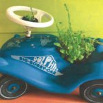 Blaues Big-Bobby-Car mit bepflanztem Anhänger vor gelbem Hintergrund