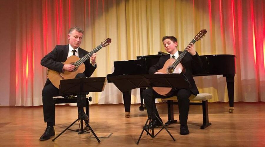 Gitarrenduo bei einem Konzertauftritt Zwei Gitarristen spielen auf einer Bühne gemeinsam klassische Gitarren; hinter ihnen stehen ein Flügel und Notenständer.