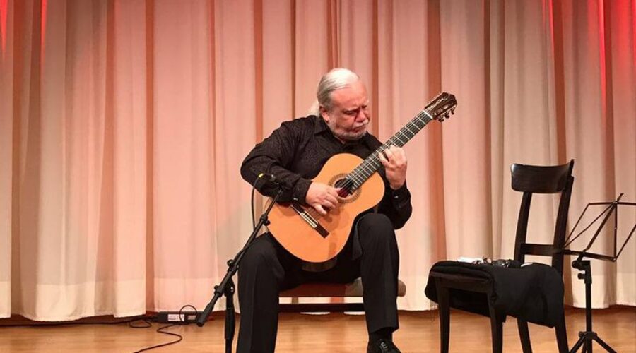 Gitarrist bei einem Konzertauftritt Älterer Gitarrist spielt eine akustische Gitarre auf einer Bühne vor einem Vorhang; daneben stehen ein Stuhl und ein Notenständer.