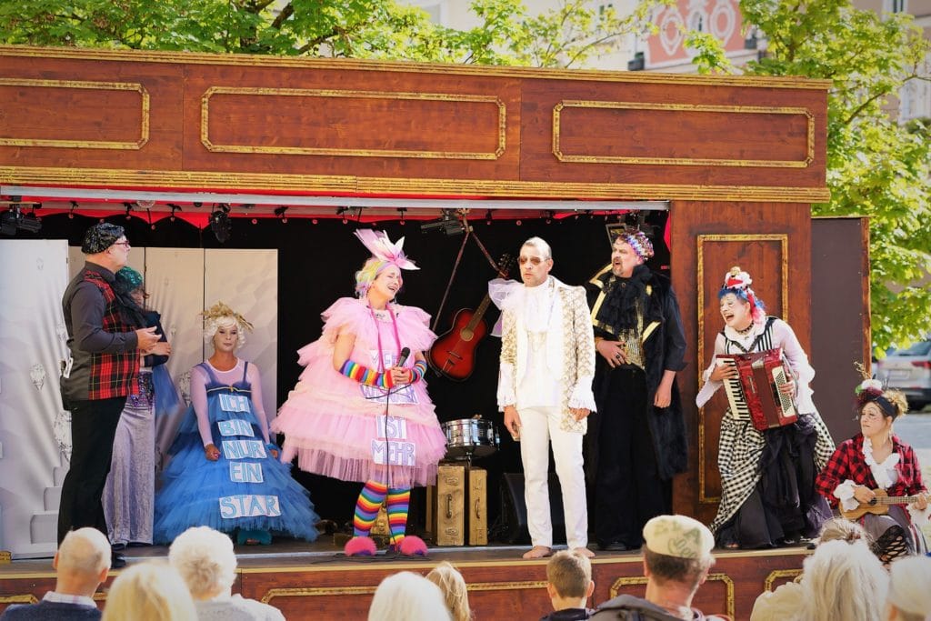 Kostümierte Schauspielerinnen und Schauspieler stehen bei einer Freiluftaufführung auf einer kleinen Holzbühne; eine Person spielt Akkordeon.