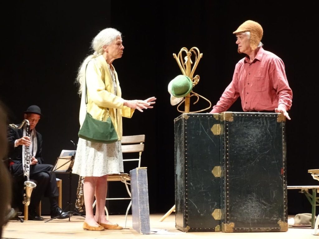 Fremd.Sein – Theateraufführung in Deggendorf Zwei Schauspielende stehen auf einer Theaterbühne neben einem großen schwarzen Koffer; links sitzt ein Musiker mit Klarinette.