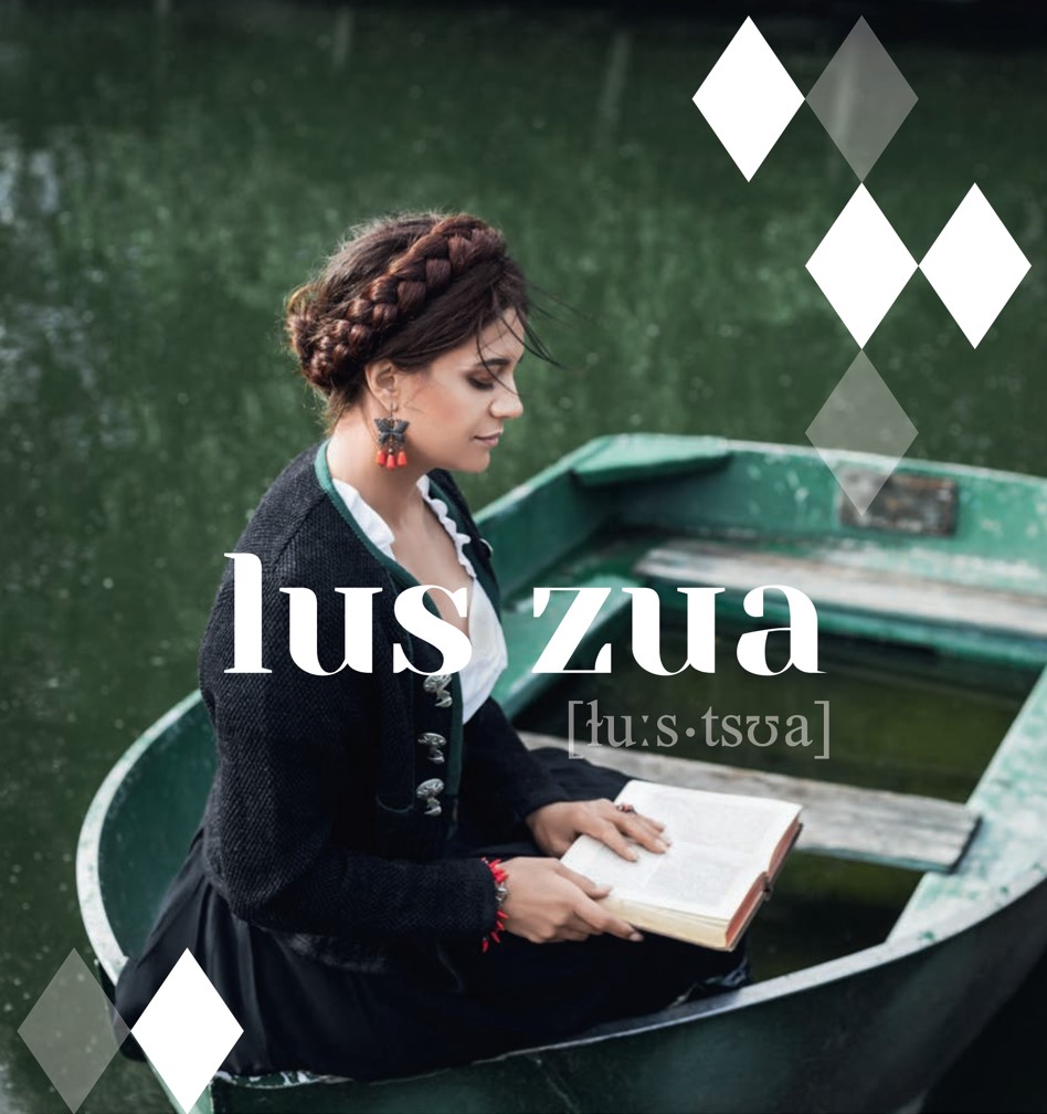 lus zua – Mundart-Veranstaltung in Deggendorf Frau in traditioneller Kleidung liest in einem Ruderboot in einem grünen Gewässer; darüber steht „lus zua“ mit phonetischer Ausspracheangabe.