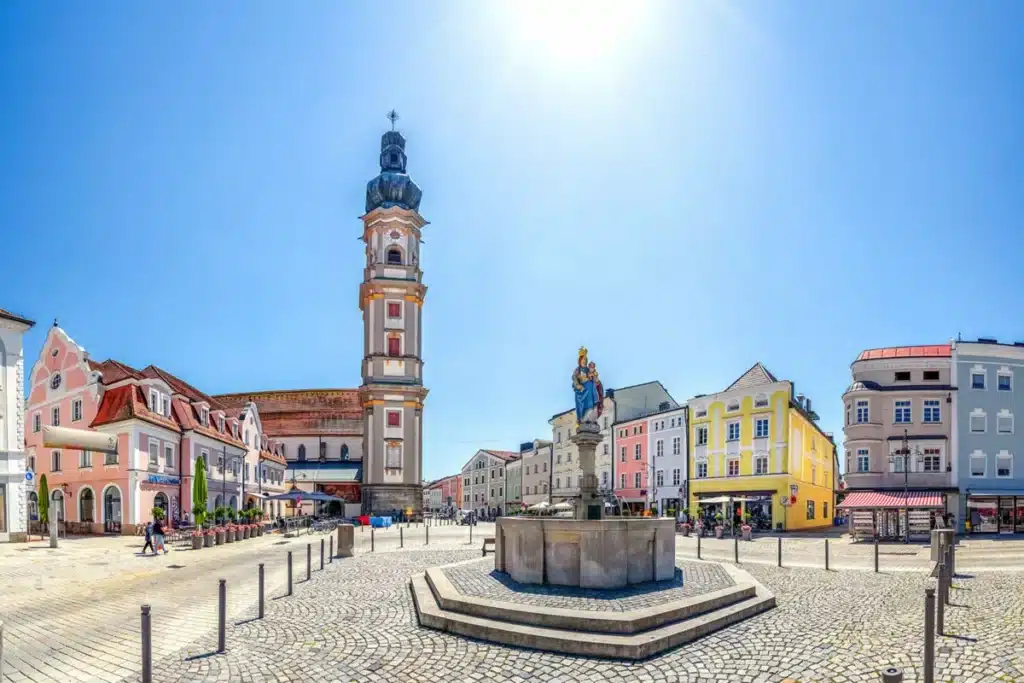 Historischer Stadtplatz in Deggendorf mit der Grabkirche, bunten Bürgerhäusern und einem Brunnen im Sonnenschein
