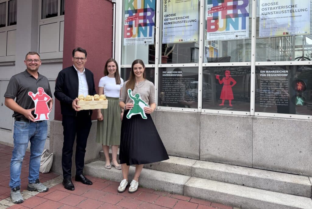 Knödelwerferin vor dem Ampelschild zur Kunstausstellung Vier Personen stehen vor einem Eingang mit Plakaten zur großen ostbayerischen Kunstausstellung; zwei halten rote und grüne Figuren auf Schildern, eine Person ein Tablett mit Knödeln.