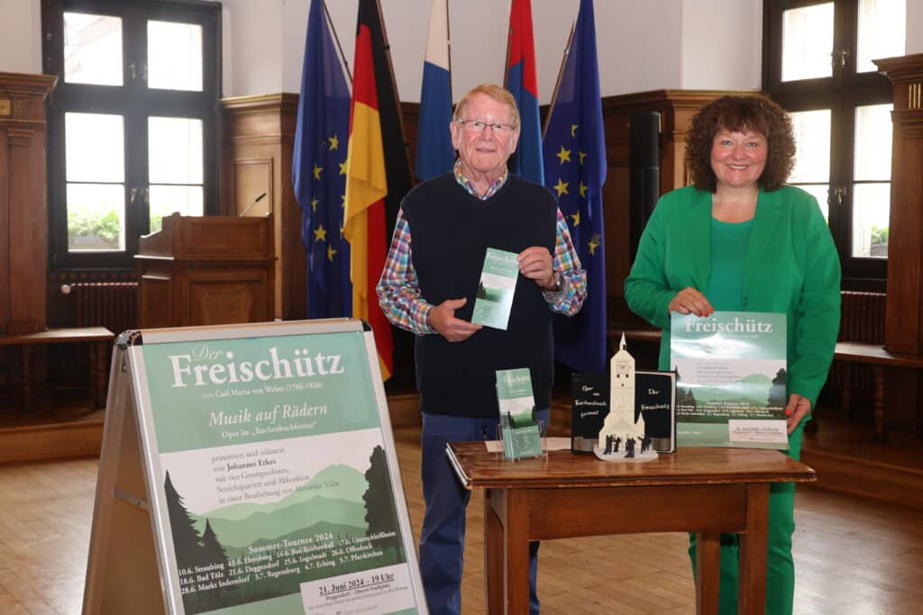 Musik auf Rädern – Oper im Taschenbuchformat Zwei Personen präsentieren Informationsmaterial zur Oper „Der Freischütz“ an einem Tisch im Rathaus; daneben steht ein Plakat zur Veranstaltung „Musik auf Rädern“.